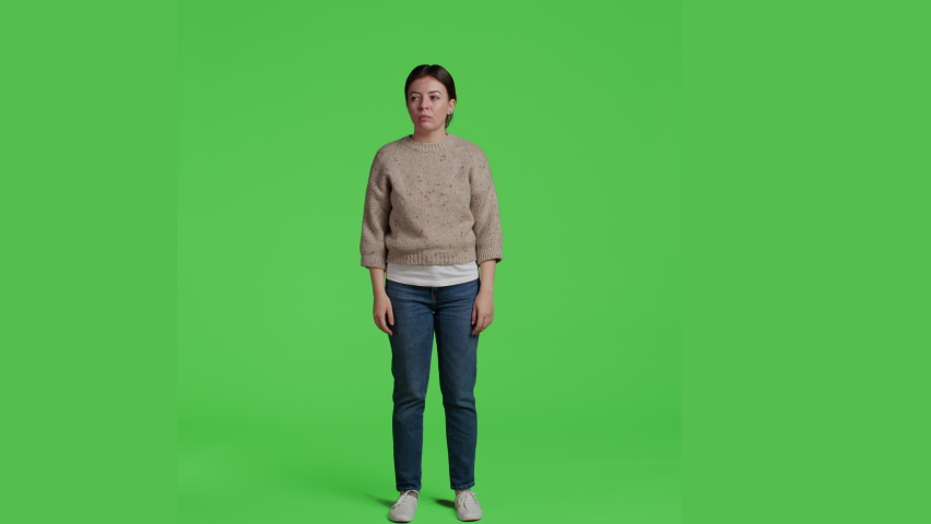 Front view of woman sitting curious and impatient in studio, posing on greenscreen backdrop. Young adult standing over isolated green screen background, being thoughtful and waiting, full body.