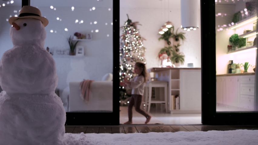 Cute baby girl opens sliding doors of cozy apartment with snowfall and snowman at the forground on Christmas holidays | Shutterstock HD Video #1097636187