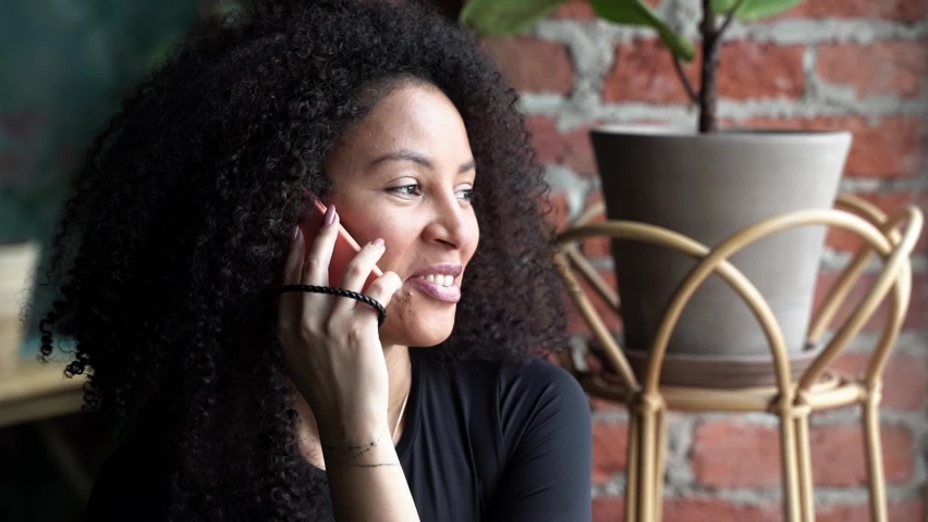 Black happy woman with African curls speaking on cellphone at home or cafe indoors. Communication with friends of relatives. Comfort relationships. Freelance | Shutterstock HD Video #1097637177
