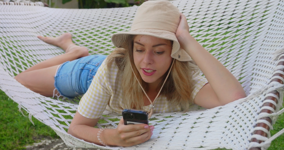 Smiling blonde woman with sunglasses using smartphone, lying relaxing on the hammock in the garden, free time and summer holiday concept to surf the internet or chat with friends using social media | Shutterstock HD Video #1097640869