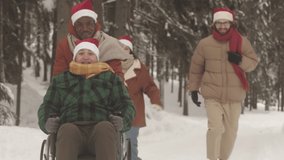 Diverse company of young cheerful people in Christmas hats having fun running along snowy pine trees in winter forest. One of women sitting in wheelchair - Powered by Shutterstock - Get 15% off with code: PIKWIZARD15