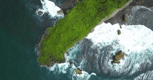 Overhead drone shot of huge coral rock overgrown by green grass that hits by the wave