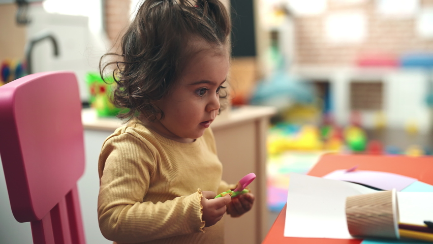 Adorable hispanic girl student holding scissors at kindergarten