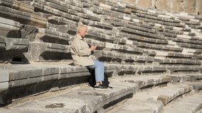 A young pretty female student is sitting on the steps of an ancient amphitheater and texting in messengers in a smartphone. Student of the Faculty of History. High quality 4k footage