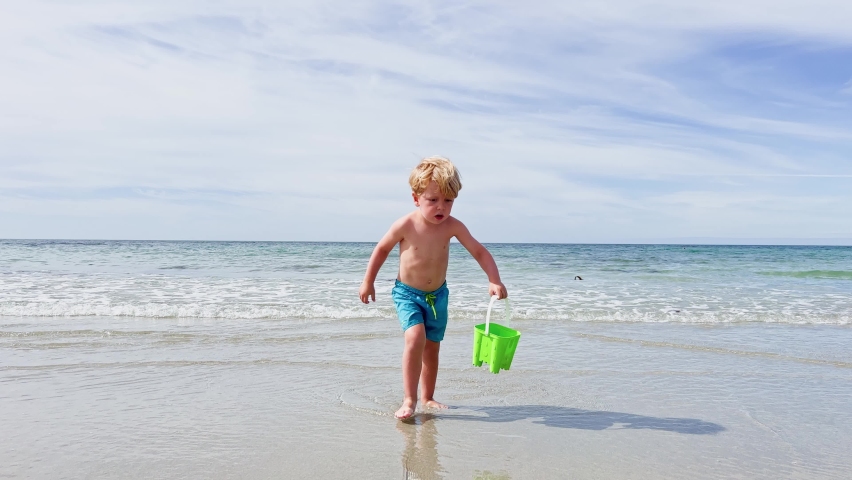 Happy little Caucasian blond boy run with plastic toy bucket on the summer beach over ocean waves and pour water on sand Royalty-Free Stock Footage #1097654677