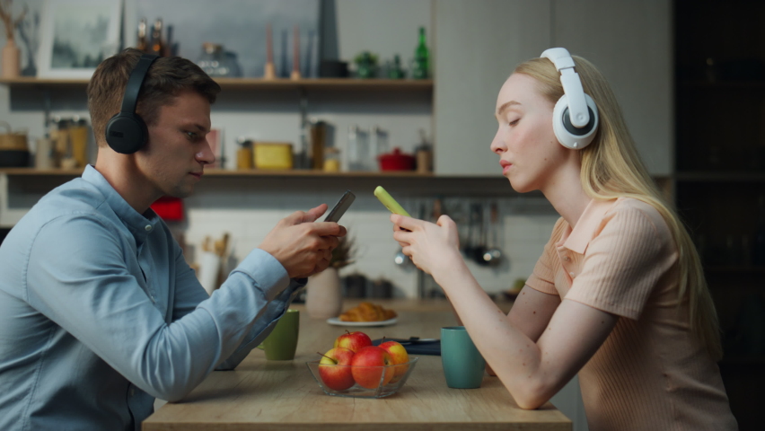 Young married couple ignoring communication addicted to modern gadgets close up. Unhappy family sitting at kitchen table listening music with headphones separately. Sad man woman feeling loneliness.