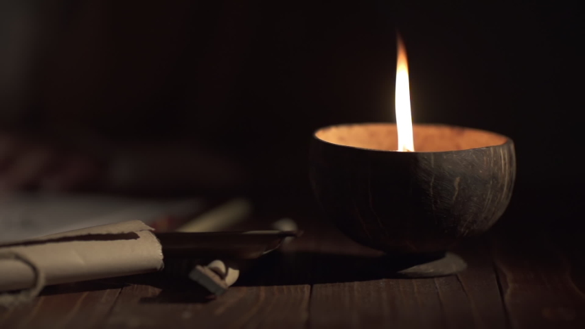 A medieval man, in out of focus, uses an old pen with ink to write Arabic script on an a paper lit by a candle,slow motion