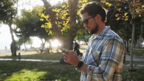 Stylish handsome young man wearing plaid shirt and sunglasses standing at the park used mobile phone, watches videos and using social networks.