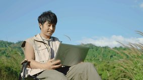 4K, Young Asian traveling man relax and enjoy outdoor lifestyle camping in forest mountain on summer holiday. Attractive male relaxing in chair while talking calling on video on laptop