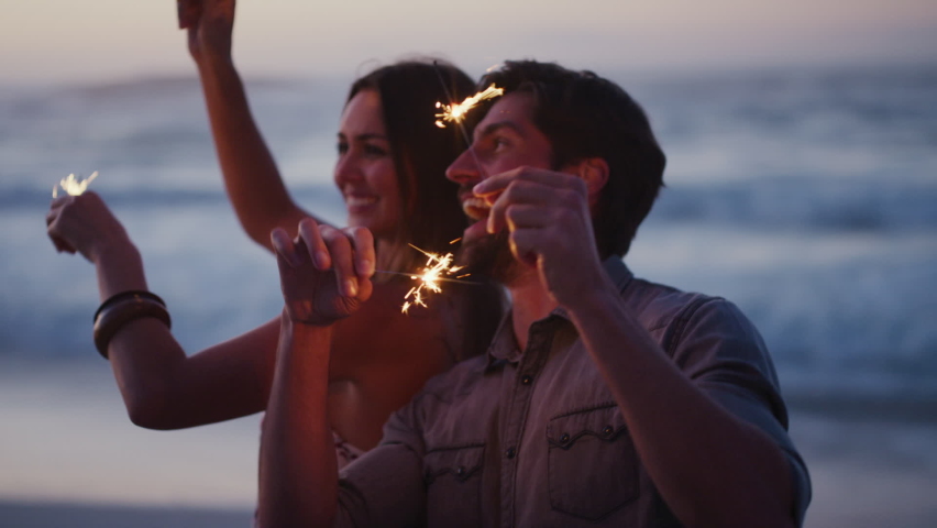 Couple, sparkler and having fun on the beach at sunset while celebrating new years eve with friends. Boyfriend and girlfriend with sparkle to celebrate on a seaside vacation or holiday in Bali Royalty-Free Stock Footage #1097691389