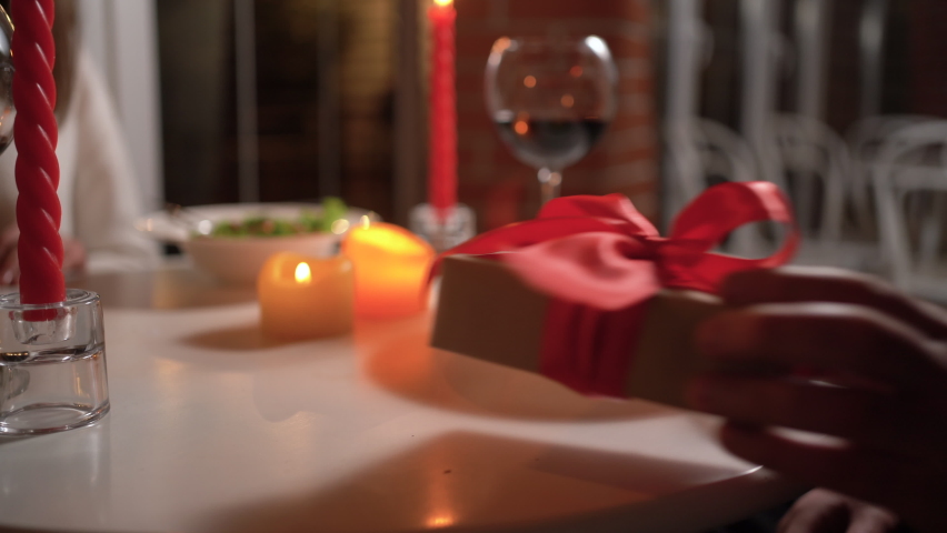 Happy Valentine's Day. Loving man giving gift to excited woman at romantic dinner in restaurant, putting it on table, greeting with 8 March holiday or anniversary, couple dating together Royalty-Free Stock Footage #1097692767