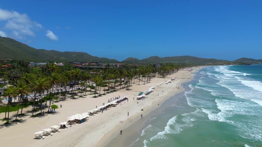 Aerial fast orbit view of the tropical Playa El Agua beach on the Margarita island. Turquoise water of the Caribbean sea, big waves and row of sun loungers on the white sand Royalty-Free Stock Footage #1097693765
