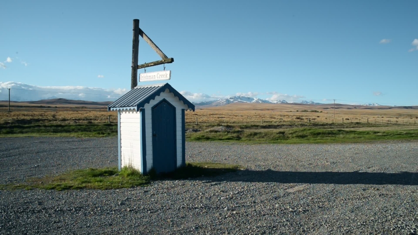 Tiny, deserted shed with a sign waving in the wind. Snow-capped mountain range and golden fields in background