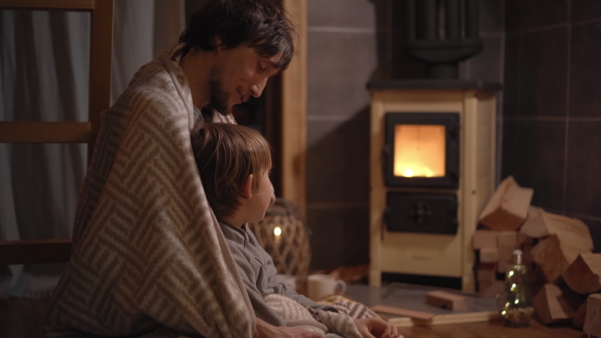 A young man and his little son wrapped in a blanket sit by a domestic stove looking at a fire. Little cozzy wooden house. Winter vacations concept