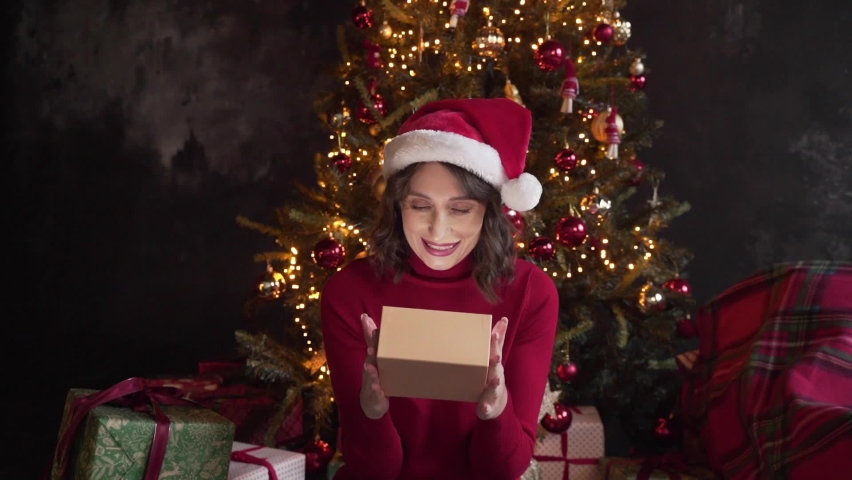 A young beautiful woman wearing red sweater and Santa's cap is opening a gift box, light shines on her face. She's happy. Christmas tree and lights on the background. Merry Xmas and Happy New Year | Shutterstock HD Video #1097712269