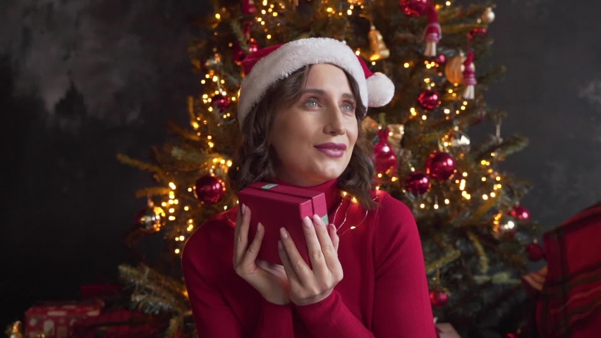 A young beautiful woman wearing red sweater and Santa's cap looks at a box with a gift, shakes it, tries to guess what is inside. Christmas tree and lights on background. New Year | Shutterstock HD Video #1097712273