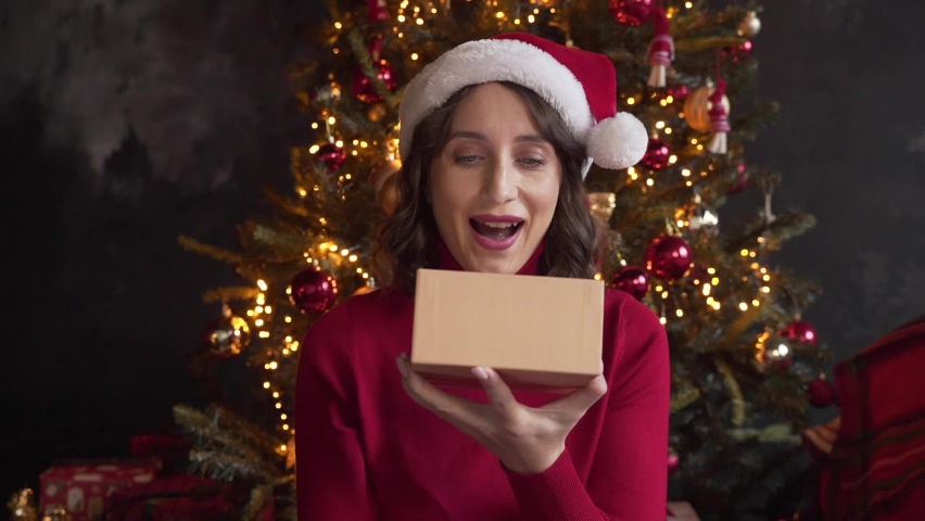 A young beautiful woman wearing red sweater and Santa's cap is opening a gift box, light shines on her face. She's happy. Christmas tree and lights on the background. Merry Xmas and Happy New Year | Shutterstock HD Video #1097712275