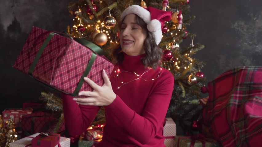 A young beautiful woman wearing red sweater and Santa's cap looks at a big box with a gift, shakes it, tries to guess what is inside. Christmas tree and lights on background. New Year | Shutterstock HD Video #1097712277