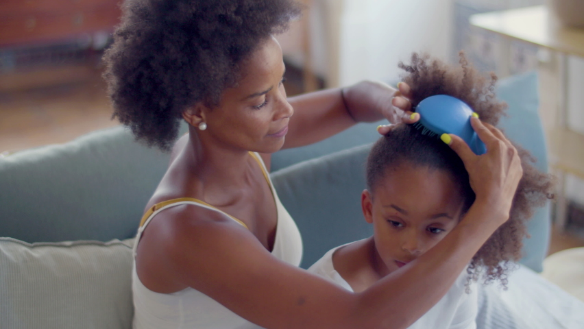 Closeup of Black woman combing daughters hair, doing ponytail sitting on couch. Short-haired mother and little cute girl talking while spending time together on lazy weekend. Family, childhood concept Royalty-Free Stock Footage #1097712697