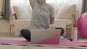 Tilt up shot of woman sitting on exercise mat, watching online yoga class on laptop and clasping hands behind back while practicing at home