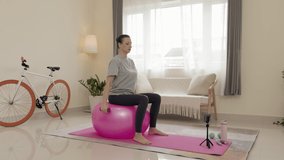 Woman sitting on exercise ball and practicing overhead tricep extension with dumbbells while doing online workout with smartphone on tripod at home