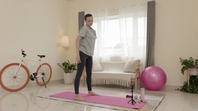 Full shot of woman doing squats and watching online video workout on smartphone held by tripod while exercising at home