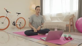 Woman with wireless earphones sitting on exercise mat at home and discussing workout program with fitness instructor via online video call on laptop