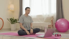 Woman with wireless earphones sitting on exercise mat, speaking with fitness coach via online web call on laptop and stretching arm while working out at home