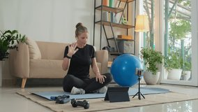 Female fitness instructor in wireless earphones waving and speaking via video call laptop while training online from home