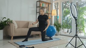 Female fitness instructor with headset explaining how to do lunge while teaching online or filming video workout with ring light and smartphone on tripod
