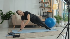Female fitness coach doing side plank on exercise mat and talking in mic while training online or filming video workout with ring light and smartphone on tripod