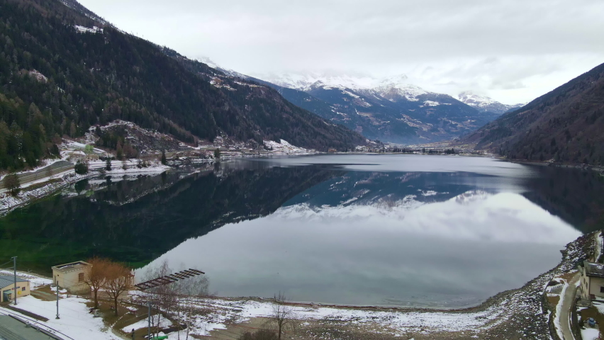 Incredible beauty of Alps mountains in winter time, drone view shot of half frozen lake located between two rock hills covered with snow. Snowy mountains reflecting in dark green lake surface in