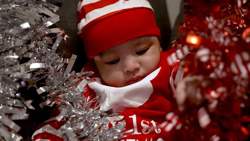 Adorable 2 Month Old Baby Boy Wearing Red Christmas Outfit And Hat And Being Playful With Red And Silver Tinsel Surrounded Him On Sofa. Slow Motion
