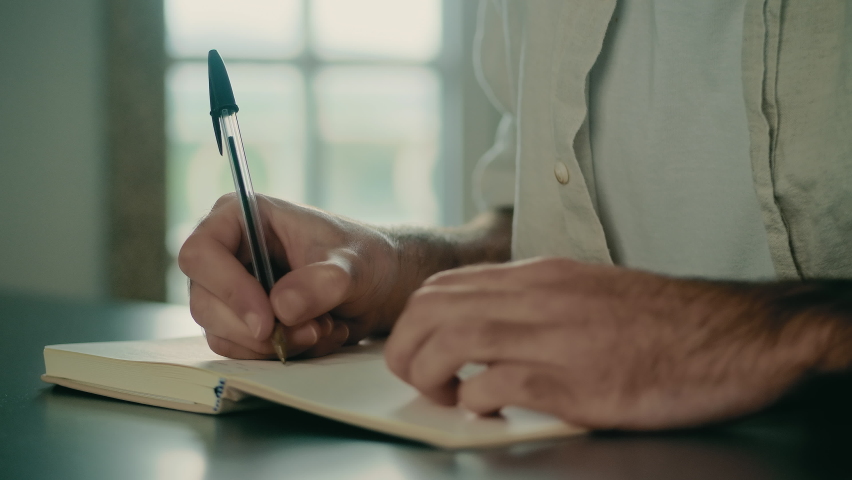 Male hands holding a pen, writing, and journaling in a small notebook. Writer, author, biographer, dramatist, journalist, novelist, screenwriter, or columnist writing in a journal. Close-up view.