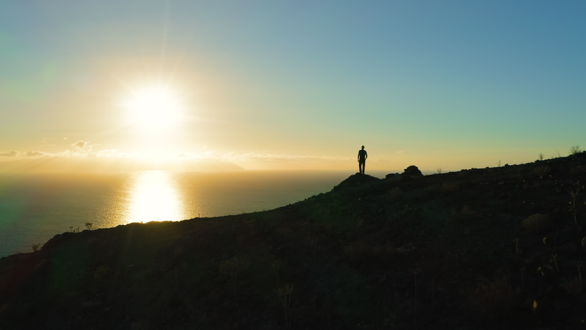 Man silhouette stands on edge of cliff and looks out at ocean during sunset. Sun touches water surface golden path in sea. Aerial drone flight. Los Gigantes Tenerife Canary Islands Spain Europe.