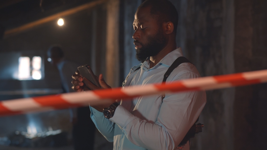 Portrait of African-American detective using smartphone at crime scene. Confident African-Americanpoliceman in suspenders checking murder details on mobile phone standing behind caution tape Royalty-Free Stock Footage #1097751285