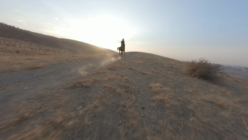 Man equestrian gallop riding on horse at sunset desert mountain suburb route enjoy freedom aerial view. FPV sports drone shot male horseman ride farm animal hilly terrain nature countryside landscape