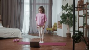 Little girl doing cardio exercise in living room at home on fitness mat at home in front of open laptop. Teenager kid doing workout looking laptop in light room interior.