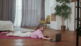 Little girl doing stretching exercises, practicing yoga on fitness mat at home in front of open laptop. Teenager kid doing horizontal scale workout looking laptop in light room interior.