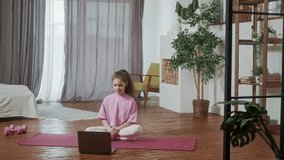 Funny adorable little girl with dark curly hair wearing pink clothing doing sport alone at home in living room in front of open laptop. Home gymnastic. Kids sport. Online training.