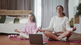 Happy hispanic woman family young healthy mom teaching cute little kid daughter meditate together in living room at home in front of open laptop , Mother and child girl do yoga exercise relax together
