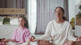Young hispanic mom and cute little kid daughter meditating in lotus pose at home, happy healthy family woman and small child girl having fun doing yoga exercise relaxing together in living room.