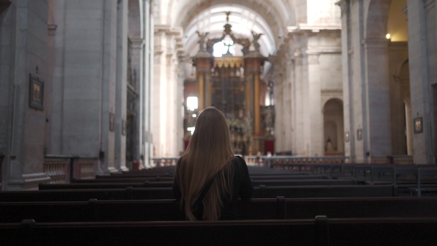 Woman with long blonde hair prays while sitting on bench inside church, rear view. Catholic Church inside evening prayer, dark mystic atmosphere female prayer sitting pew at Church religion concept | Shutterstock HD Video #1097763265