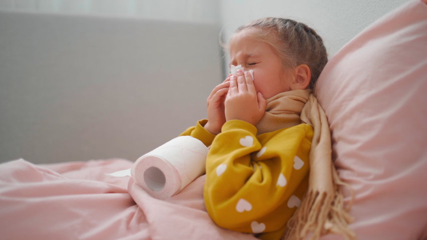 Child girl in plaid and scarf sneezes snot into napkin on sofa at home Child cold flu illness tissue blowing runny nose. toddler girl is lying in bed and blowing nose into tissue paper at home