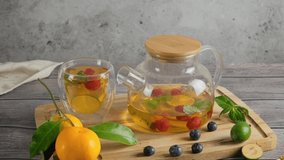 Woman taking a cup with green refreshing tea flavored with fruit, berries and mint, transparent glass cup and teapot with brewing tea placed on wooden table, slow motion 4k video footage