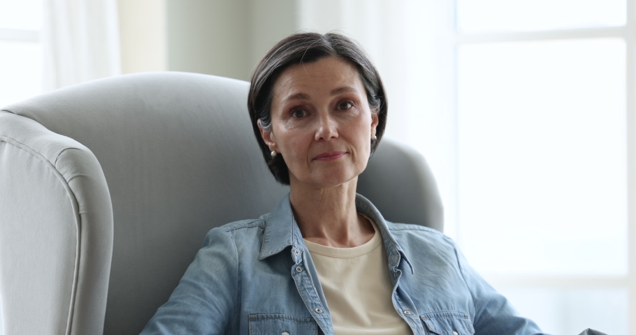 Head shot of attractive middle-aged woman smile look at camera relaxing alone seated on cozy armchair at home feels happy, enjoy weekend leisure at own or rented apartment. Carefree retiree portrait Royalty-Free Stock Footage #1097784883
