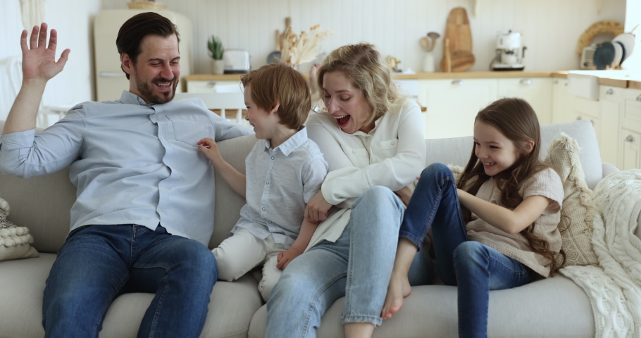 Happy playful family tickling each other seated on couch at home. Carefree parents play with beautiful little son and daughter enjoy funny weekend leisure together at modern cozy own house. Playtime Royalty-Free Stock Footage #1097784929