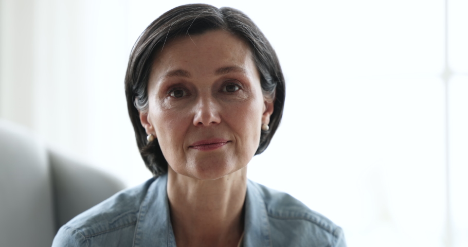 Close up attractive brunette middle-aged 55s female smile staring at camera seated indoor. Portrait of happy retiree woman spend time alone at home, having pleasant appearance, looks good, enjoy rest Royalty-Free Stock Footage #1097785083