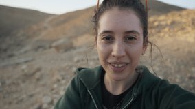 Female hiker or media content creator has video call on mobile in the desert. Young woman influencer or traveler having conversation on smartphone outdoors. Smiles and happy waving at camera on a trip