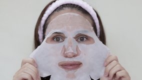woman face portrait with facial textile paper sheet anti wrinkle aging moisturizing mask.girl applying sheet with hands brunette hair pink headband slow motion video.face skin care millenial woman